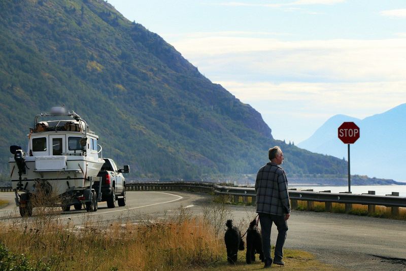 A pickup tows a cabin boat along the water in Alaska while a man walks two dogs.