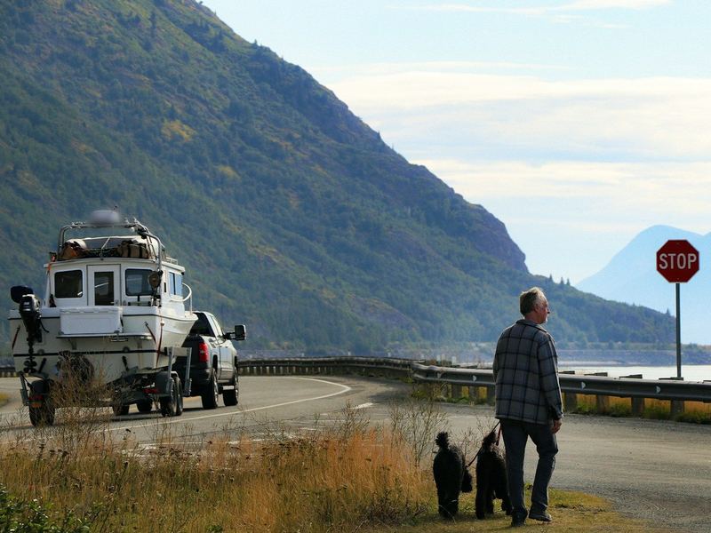 A pickup tows a cabin boat along the water in Alaska while a man walks two dogs.
