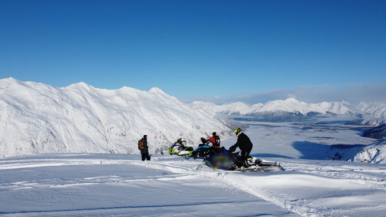 Snowmachiners stop on a snowy ridge above a mountain valley in Alaska.